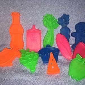 May include: A set of 12 colorful plastic cookie cutters shaped like various foods, including a bottle of soda, a bunch of grapes, a cupcake, a slice of pizza, a celery stalk, a carrot, an apple, a fish, a gingerbread man, and a loaf of bread.