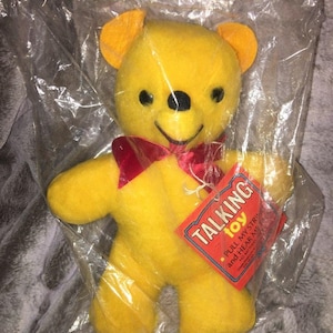 Puede incluir: Un oso de peluche amarillo con una pajarita roja. El oso está empaquetado en una bolsa de plástico transparente con una etiqueta roja y blanca que dice "Talking Toy - Pull my string and hear me talk".