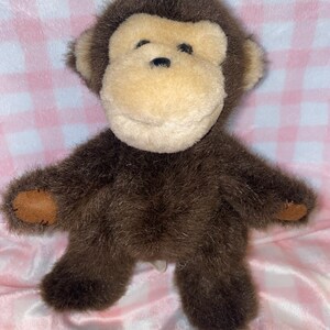 May include: A plush monkey toy with brown fur, a tan face, and brown accents on the ears and feet. The stuffed animal has a smiling expression and is sitting on a pink and white checkered surface.