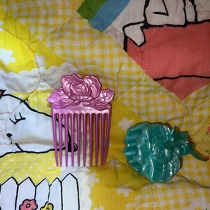 May include: A close-up of two plastic hair accessories: a pink comb with a rose design and a teal-colored flower-shaped clip. The items are on a yellow patterned surface.