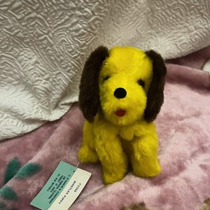 Puede incluir: Un juguete de peluche amarillo de Whistler Puppy con orejas marrones, nariz negra y boca roja. El juguete está sobre una manta rosa. La etiqueta dice "Whistler Puppy" y "C.R. Dakin & Company."