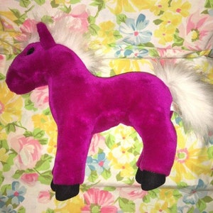 Puede incluir: Un unicornio de peluche de color magenta vibrante. Cuenta con una melena y cola blancas, cascos negros y un solo cuerno. El juguete está diseñado en posición de pie, mostrando sus detalles.