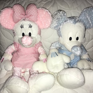 Op de afbeelding: Een paar pluche Mickey Mouse en Minnie Mouse speelgoed. Minnie Mouse draagt een roze en wit geruit jurkje met een roze strik. Mickey Mouse draagt een blauw en wit geruit overhemd. Beide speelgoeddieren hebben witte vacht en zitten op een wit gestikt oppervlak.