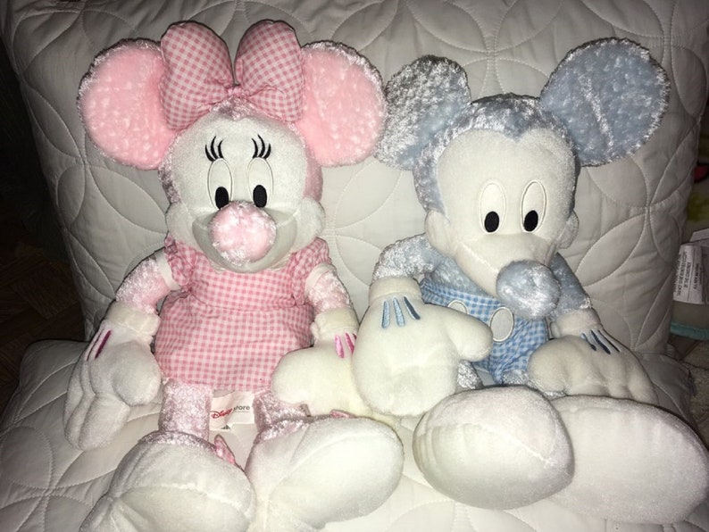 Op de afbeelding: Een paar pluche Mickey Mouse en Minnie Mouse speelgoed. Minnie Mouse draagt een roze en wit geruit jurkje met een roze strik. Mickey Mouse draagt een blauw en wit geruit overhemd. Beide speelgoeddieren hebben witte vacht en zitten op een wit gestikt oppervlak.