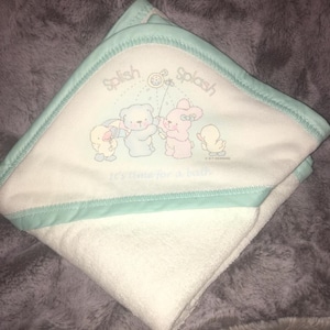 Puede incluir: Toalla con capucha blanca para bebé con ribete azul claro. La toalla presenta una ilustración de dibujos animados de un pato, un oso y un conejito con las palabras "Splish Splash" y "It's time for a bath".