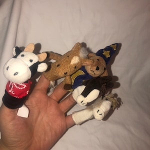 May include: Four finger puppets, a cow, a horse, a bear wearing a wizard hat, and a horse, are worn on a person's fingers. The puppets are made of soft, plush fabric.