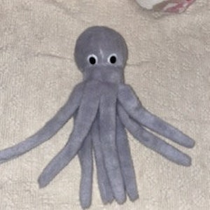 May include: A gray plush octopus toy with eight long tentacles and two googly eyes. The stuffed animal is made of soft fabric and is sitting on a white surface with a floral pattern.