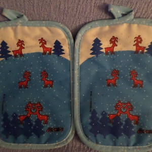 May include: Two quilted pot holders with a winter design. They feature red deer, blue evergreen trees, and a light blue background with white polka dots. Each pot holder has a hanging loop. Made of fabric.