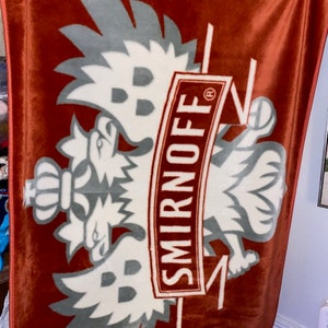 May include: A rust-colored blanket featuring a large, stylized emblem with a double-headed eagle design in white and gray. The word "SMIRNOFF" is prominently displayed in a rectangular frame. The blanket is hanging from clothespins.