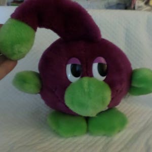 Puede incluir: Un juguete de peluche morado y verde con ojos grandes y un cuello largo y curvo. El juguete tiene un cuerpo redondo y pies verdes.
