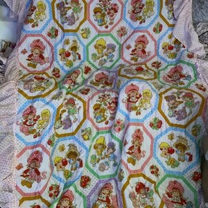 Pode incluir: Um edredom vintage com um padrão repetido de personagens da Moranguinho em molduras hexagonais coloridas. O edredom tem um fundo branco com detalhes em rosa, verde, azul e amarelo. As bordas são adornadas com uma babado de bolinhas rosa e branco.