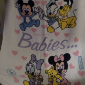 Puede incluir: Manta de bebé blanca con corazones rosas y personajes de dibujos animados, incluyendo Mickey Mouse, Minnie Mouse, Donald Duck y Goofy. El texto "Only for Babies..." está impreso en morado.