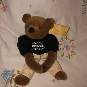 Puede incluir: Un oso de peluche marrón con una camiseta negra con el texto "KNOCK, BEFORE ENTERING". El oso tiene pantalones color beige y una etiqueta "RUSS". El peluche está sobre una superficie con estampado floral.