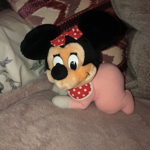 May include: A pink and black plush toy of a baby Minnie Mouse wearing a red and white polka dot bib. The toy is lying on a pink fuzzy blanket.