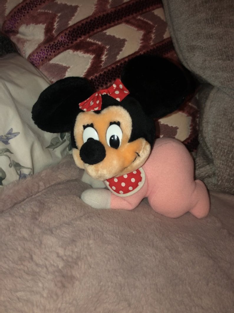 May include: A pink and black plush toy of a baby Minnie Mouse wearing a red and white polka dot bib. The toy is lying on a pink fuzzy blanket.