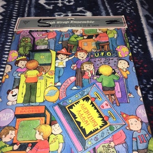 May include: A vintage gift wrap ensemble featuring a blue background with colorful illustrations of children playing arcade games. The wrap includes images of classic arcade games like "Space Invaders", "Cosmic Raiders", "The Thing", and "U.F.O." The text "Happy Birthday" is featured on a yellow background.