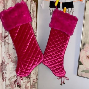 May include: Two pink quilted Christmas stockings with faux fur trim and silver bells hanging from the bottom.