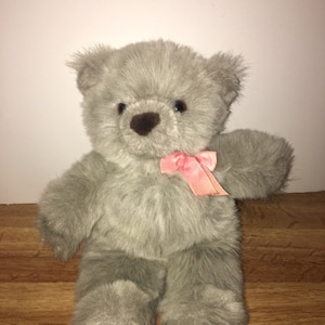 Könnte beinhalten: Ein grauer Teddybär mit einer rosa Schleife um den Hals. Der Bär hat eine braune Nase und dunkle Augen. Das Fell ist weich und flauschig. Der Bär sitzt aufrecht.