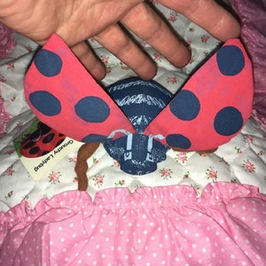 May include: A red and blue ladybug plush toy with black polka dots on its wings. The toy is sitting on a pink and white quilted fabric with a tag that reads "Grouchly Ladybug".