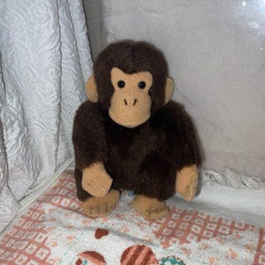 Könnte beinhalten: Ein Stofftier-Schimpanse mit braunem Fell und beigen Gesichtszügen. Das Plüschtier hat schwarze Augen und sitzt aufrecht. Das Spielzeug befindet sich auf einer gemusterten Oberfläche mit einem strukturierten weißen Hintergrund.