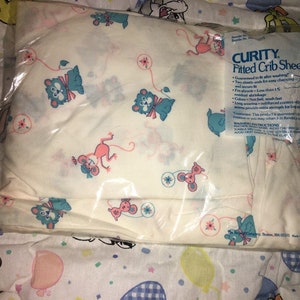 May include: A Curity fitted crib sheet in a clear plastic package. The sheet is white with a repeating pattern of blue bears and pink mice. The package has the text "CURITY Fitted Crib Sheet".