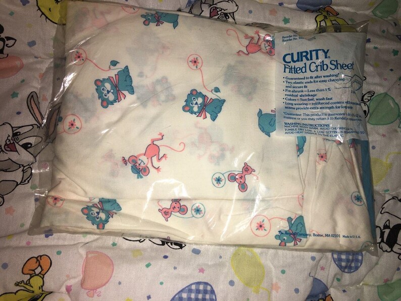 May include: A Curity fitted crib sheet in a clear plastic package. The sheet is white with a repeating pattern of blue bears and pink mice. The package has the text "CURITY Fitted Crib Sheet".