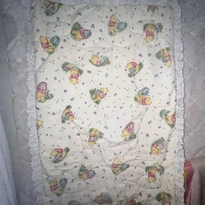 May include: A white quilted baby blanket with a white lace border. The blanket features a repeating pattern of Winnie the Pooh characters in various poses.