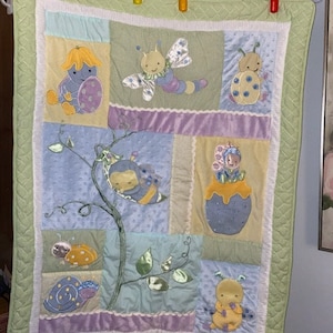 May include: A multi-colored baby quilt with a patchwork design featuring various embroidered insects and flowers. The quilt has a green border and is made of soft, plush fabric.