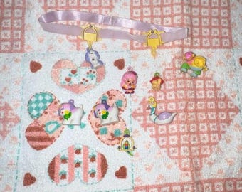 May include: A collection of small, colorful, plastic figurines on a white and pink patterned background. The figurines include a swan, a unicorn, a dog, a fairy, a bird, and a flower. There is also a pink ribbon with a gold charm.