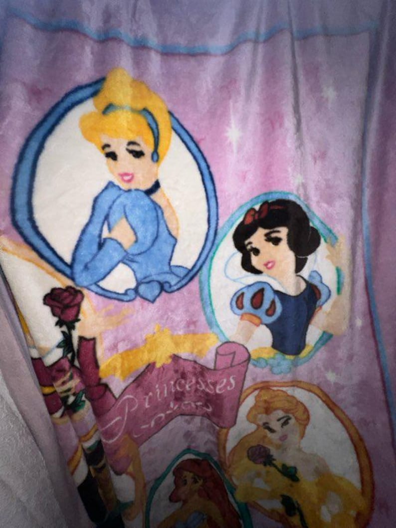 K&ouml;nnte beinhalten: Eine rosa Fleece-Decke mit einem Design, das Disney-Prinzessinnen zeigt. Die Prinzessinnen sind Cinderella, Schneewittchen, Belle und Arielle. Die Decke hat den Text "Princesses" darauf.