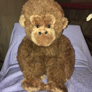 May include: A brown plush gorilla toy with black eyes and a brown nose. The toy is sitting on a purple blanket.