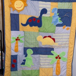 May include: A blue and yellow patchwork baby quilt with a sun, palm trees, and dinosaurs. The quilt is made of different fabrics and has a quilted design.