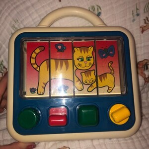 May include: A white plastic toy with a blue frame and a handle. The toy has a sliding puzzle with a picture of a mother cat and a kitten. The puzzle has three buttons on the bottom, one green, one red, and one yellow.