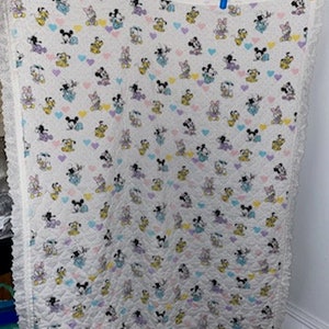 May include: White baby quilt with a repeating pattern of Mickey Mouse, Minnie Mouse, Donald Duck, and other Disney characters, along with pastel hearts. The quilt has a ruffled edge and is hanging from clothespins.