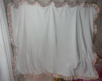 May include: White baby blanket with a ruffled edge. The ruffle is decorated with a floral pattern in pink, yellow, and green. The blanket is hanging, showing its soft texture and design.