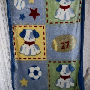 May include: A baby blanket with a blue border and a patchwork design. The blanket features squares with images of a baseball, stars, a soccer ball, a rugby ball with the number 27, and cartoon dogs.