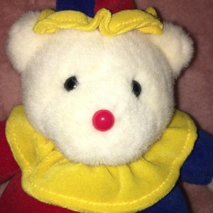 Op de afbeelding: Een witte pluche teddybeer met een rode, blauwe en gele narrenmuts en een gele en rode kraag. De beer heeft een rode knoopneus.