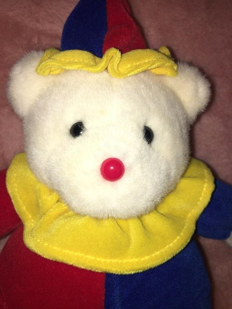 Op de afbeelding: Een witte pluche teddybeer met een rode, blauwe en gele narrenmuts en een gele en rode kraag. De beer heeft een rode knoopneus.