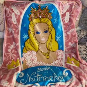 Puede incluir: Manta de felpa rosa con una ilustración de Barbie de "Barbie y el Cascanueces". El diseño incluye un retrato de Barbie con una corona, con el título de la película en una pancarta azul. Elementos decorativos: coronas y zapatillas de ballet.