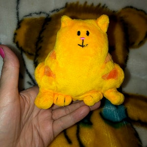 May include: A yellow plush cat toy with orange spots and a pink nose. The cat is sitting on a hand.