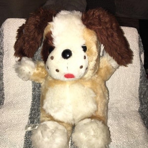 May include: A brown and white plush dog toy with floppy ears. The dog has black eyes and a red tongue. The toy is sitting on a striped blanket.