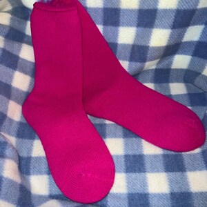 May include: A pair of vibrant fuchsia socks. The socks are made of a soft material and are displayed on a blue and white checkered blanket. The socks are a solid color and appear to be new.