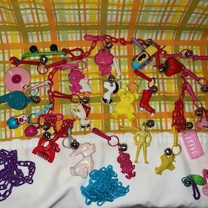 May include: A collection of colorful vintage keychains featuring various shapes and designs. Includes a pink frog, a yellow bear, a red roller skate, a pink bicycle, and other novelty charms. The keychains are attached to red chains.