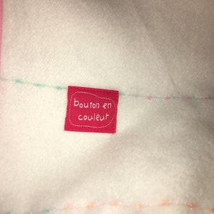 Peut inclure: Gros plan sur un tissu blanc avec une bordure rose et une petite étiquette rouge. L'étiquette porte les mots "bouton en couleur" en blanc. Des fils colorés sont cousus sur le tissu.