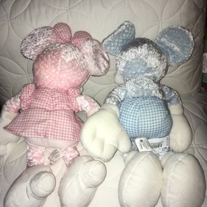 Op de afbeelding: Twee pluche speelgoedjes, een roze en een blauwe, liggen op een wit gestikt oppervlak. Beide speelgoedjes zijn in de vorm van Mickey Mouse en hebben een ruitjespatroon op hun kleding.
