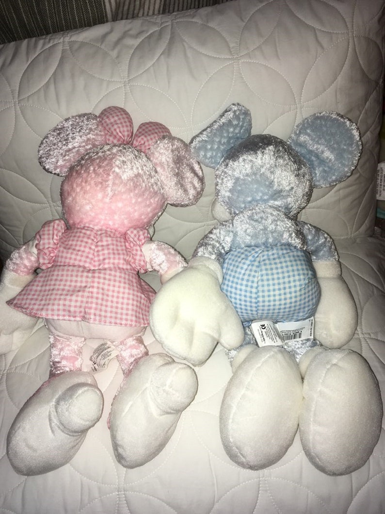 Op de afbeelding: Twee pluche speelgoedjes, een roze en een blauwe, liggen op een wit gestikt oppervlak. Beide speelgoedjes zijn in de vorm van Mickey Mouse en hebben een ruitjespatroon op hun kleding.