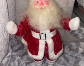 May include: A Santa Claus figurine with a red velvet suit, white trim, and a matching hat. The figure has a long white beard, rosy cheeks, and white gloves. The belt is white with a silver buckle. The figurine is approximately 12 inches tall.