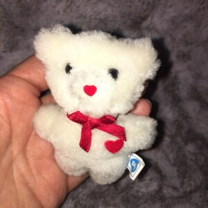 May include: A small, white teddy bear with black eyes and a red heart-shaped nose. It has a red ribbon bow and a small red heart on its chest. A tag is attached to the bear.