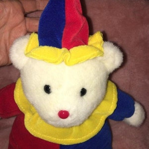 Op de afbeelding: Een witte pluche teddybeer met een rode, gele en blauwe narrenmuts en een bijpassende outfit. De beer heeft zwarte ogen en een rode neus.