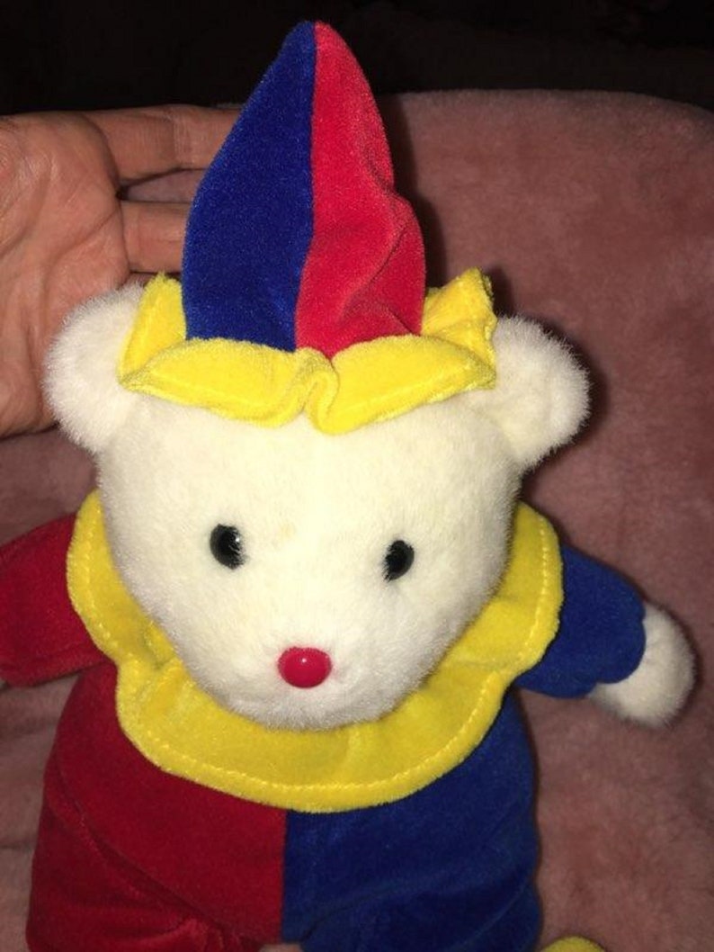 Op de afbeelding: Een witte pluche teddybeer met een rode, gele en blauwe narrenmuts en een bijpassende outfit. De beer heeft zwarte ogen en een rode neus.